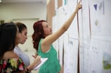 Three women are closely examining several large sheets of paper on a wall, possibly in an educational or professional setting. One woman points to a section on the paper, while another makes notes on a notepad.