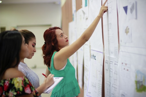 Three women are closely examining several large sheets of paper on a wall, possibly in an educational or professional setting. One woman points to a section on the paper, while another makes notes on a notepad.