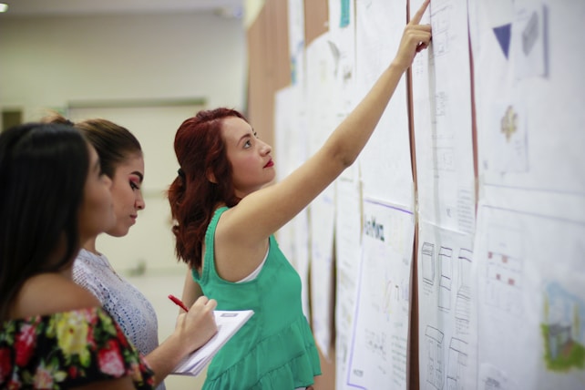 Three women are closely examining several large sheets of paper on a wall, possibly in an educational or professional setting. One woman points to a section on the paper, while another makes notes on a notepad.