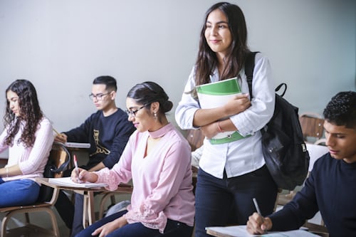 estudantes em uma sala de aula, com uma garota em pé carregando um caderno verde