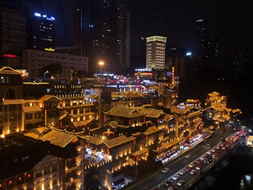 A nighttime cityscape featuring a bustling urban area with traditional Chinese architectural elements. Buildings are illuminated with warm lights, creating a vibrant contrast against the dark sky. The scene includes a busy street with a line of cars, highlighting the lively atmosphere.