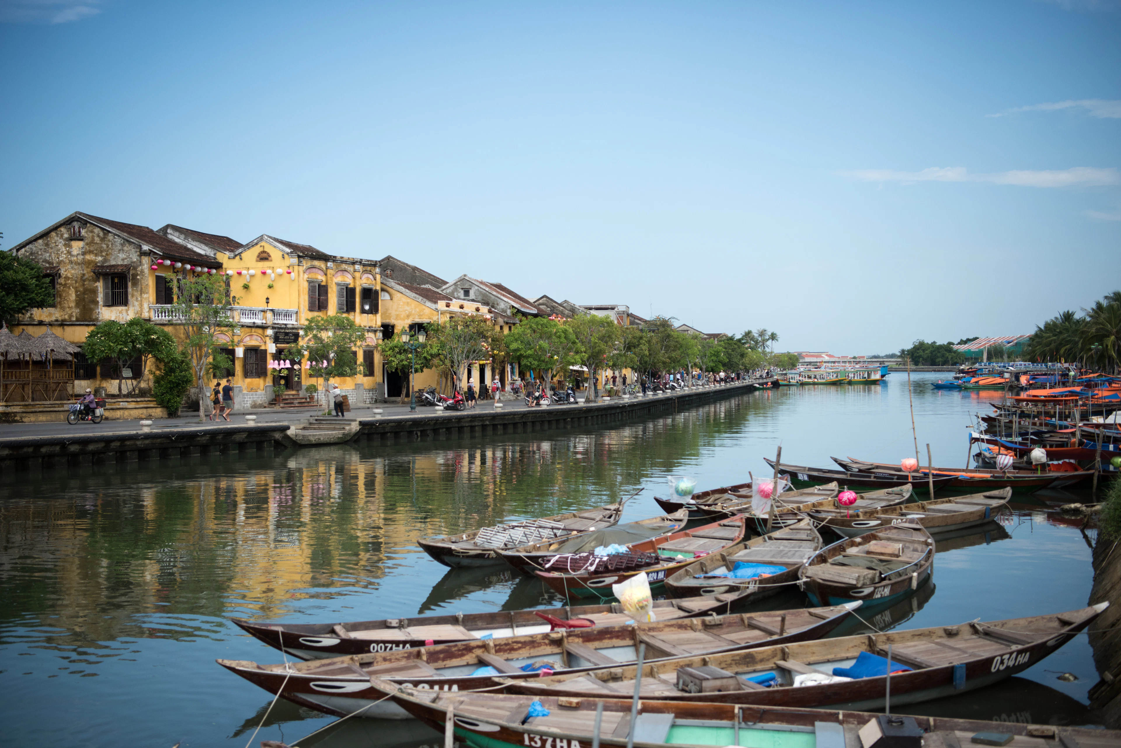 Wooden fishing boats moored along the Thu Bon river in Hoi An, with the ochre-yellow shophouses and tiled roofs of the Ancient Town lining the far bank under a clear sky