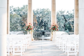 An outdoor setting is arranged for a formal event, with two columns framing red and white floral arrangements. Rows of white chairs are set up facing a small fountain and a body of water, surrounded by lush trees draped with Spanish moss.