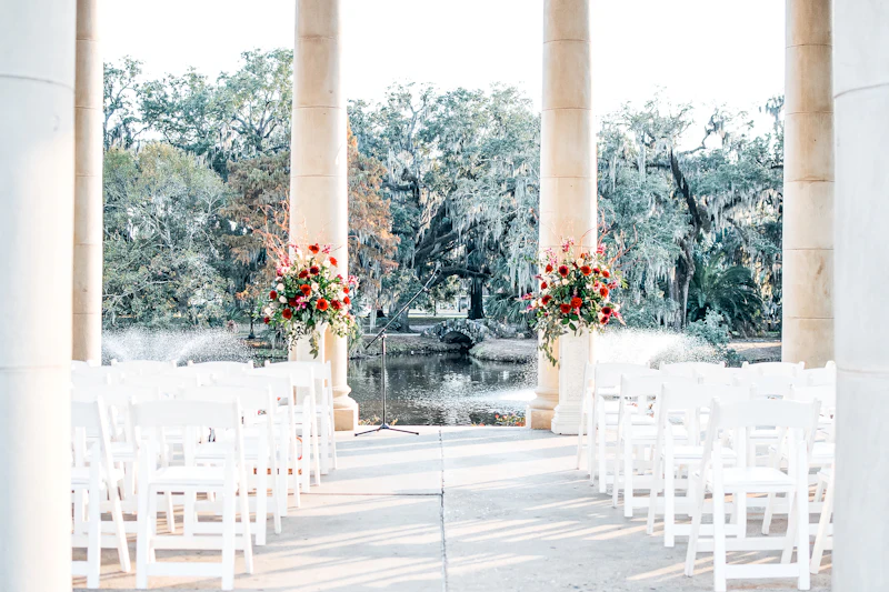 White chairs and tall floral arrangements set up for an outdoor wedding ceremony beneath stone columns