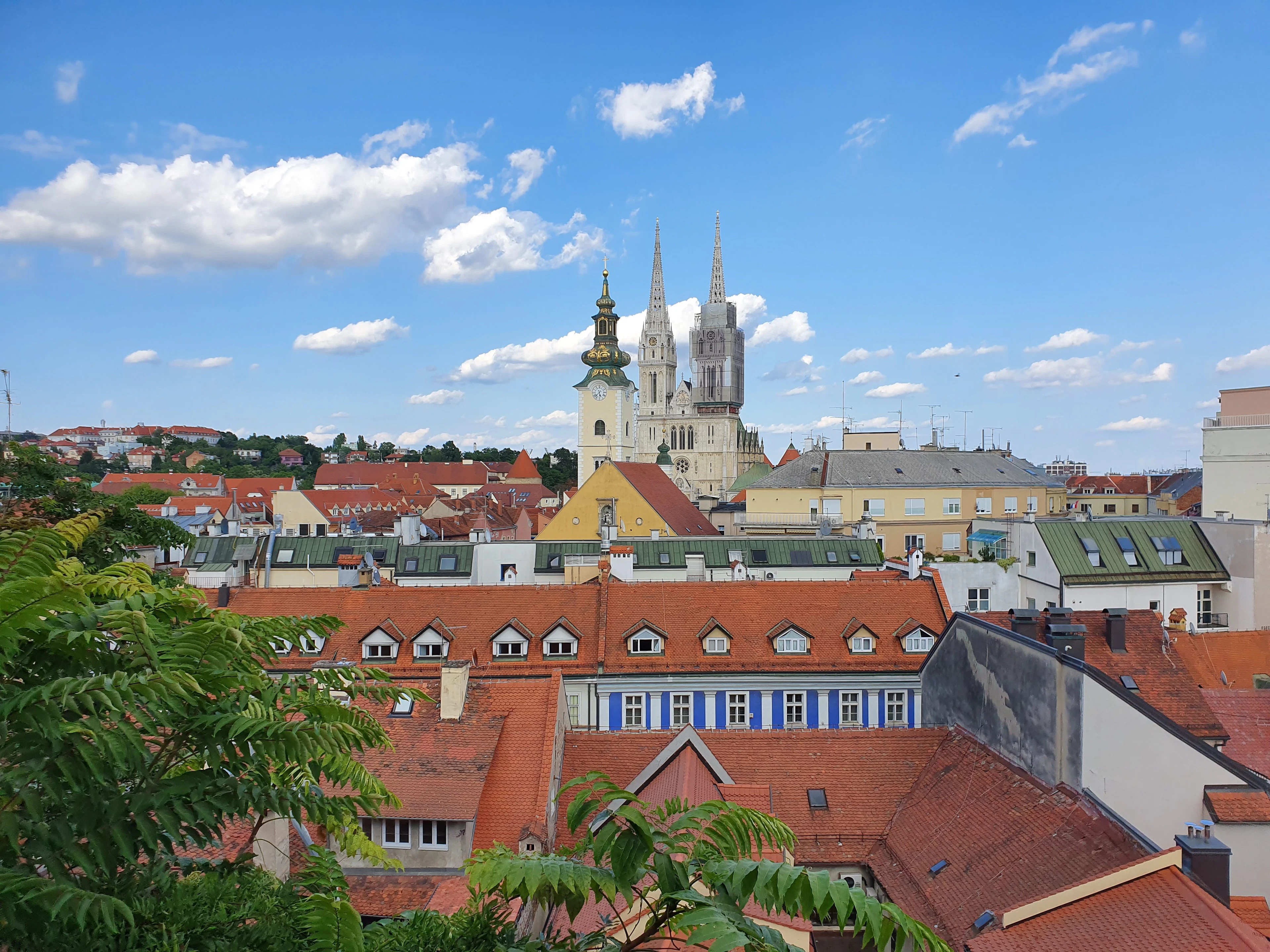 Twin cathedral spires rise above red-tiled rooftops under a blue sky, framed by green branches.