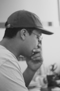 A person in profile view is wearing a cap and glasses and appears to be deep in thought, with hand resting on their chin. The background is blurred, featuring indistinct objects possibly including a cup and some stationary items.