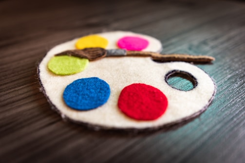 A felt art palette with five colorful felt circles representing paint, including yellow, pink, green, blue, and red. A small felt paintbrush lies across the palette.