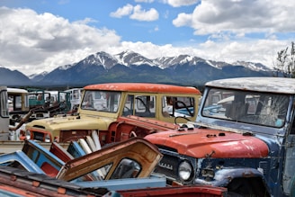 A collection of old, worn vehicles with chipped paint and broken windows, arranged in a lot against the backdrop of snow-capped mountains and a partly cloudy sky.