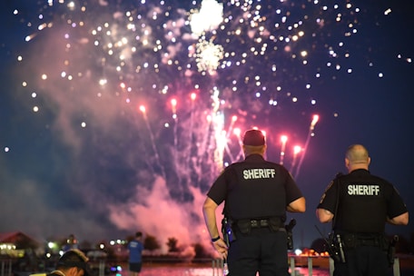 Two law enforcement officers stand by a waterfront watching a vibrant fireworks display in the night sky. The fireworks illuminate a dark, clear sky with vivid colors.