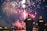 Two law enforcement officers stand by a waterfront watching a vibrant fireworks display in the night sky. The fireworks illuminate a dark, clear sky with vivid colors.