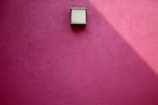 A smooth, pink plaster wall features a minimalist, rectangular light fixture casting a slanted shadow.