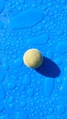 A tennis ball rests on a bright blue surface covered with water droplets, casting a shadow to the right. The surface features variously shaped droplets that reflect light, adding texture to the image.