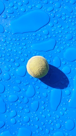A tennis ball rests on a bright blue surface covered with water droplets, casting a shadow to the right. The surface features variously shaped droplets that reflect light, adding texture to the image.