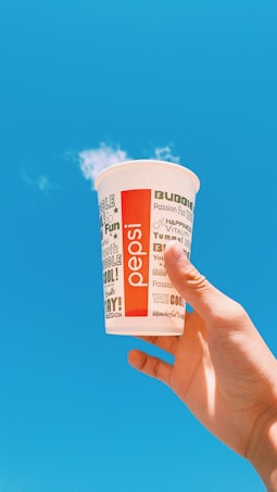 A hand holding a paper cup with Pepsi branding against a bright blue sky. The cup features various playful words like 'Passion', 'Cool', and 'Fun' in different fonts and sizes, with a red vertical stripe that says 'pepsi'.