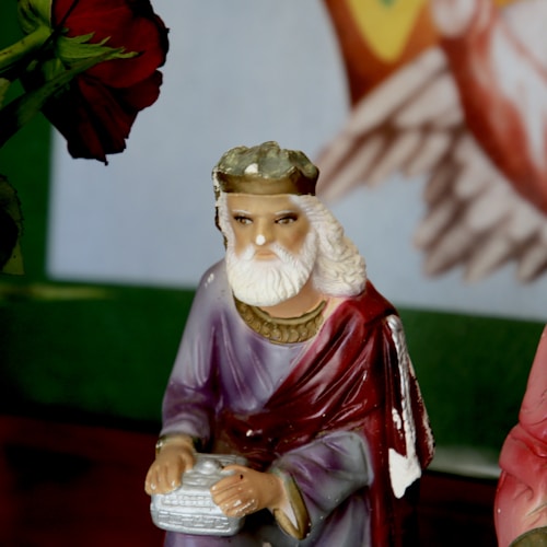 A ceramic figurine of a bearded man wearing a crown and a robe, holding a rectangular object in both hands. The figure is set against a colorful background with parts of a rose and a blurred artwork visible. The overall setting gives a sense of stillness and focus.
