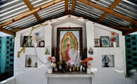 An outdoor altar with a prominent framed image of Our Lady of Guadalupe surrounded by various religious icons and statues. The altar is white and has multiple shelves with flowers, candles, and small figurines. A garland and string lights are draped across the structure. The background features a striped metal roof and brick walls.