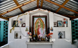 An outdoor altar with a prominent framed image of Our Lady of Guadalupe surrounded by various religious icons and statues. The altar is white and has multiple shelves with flowers, candles, and small figurines. A garland and string lights are draped across the structure. The background features a striped metal roof and brick walls.