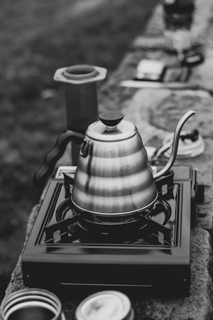 A metal kettle with a curved spout is placed on a portable gas burner. The kettle and stove are situated on a stone surface, and other camping-related items are blurred in the background. The setting appears to be outdoors.
