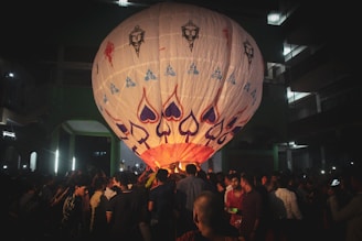 A large decorated hot air balloon is in the process of being launched in the midst of a crowded outdoor setting at night. The balloon features various vibrant designs including spade symbols and artistic faces. Numerous people are gathered around it, some assisting with its launch while others look on.