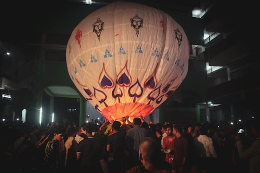A large decorated hot air balloon is in the process of being launched in the midst of a crowded outdoor setting at night. The balloon features various vibrant designs including spade symbols and artistic faces. Numerous people are gathered around it, some assisting with its launch while others look on.
