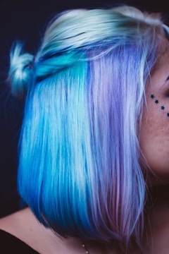Vibrant hair dyed in shades of blue and purple, styled in a bob cut. A small portion is tied in a bun, and star-shaped black face tattoos are visible on the cheek.
