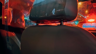 The image features the interior of a vehicle, showcasing the seat headrest and a protective partition separating the driver from the passenger area. Reflections of red and blue lights from outside the vehicle create a vibrant, urban atmosphere. There are signs with text in another language visible on the partition.