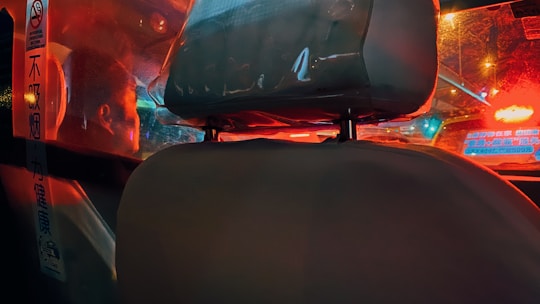 The image features the interior of a vehicle, showcasing the seat headrest and a protective partition separating the driver from the passenger area. Reflections of red and blue lights from outside the vehicle create a vibrant, urban atmosphere. There are signs with text in another language visible on the partition.