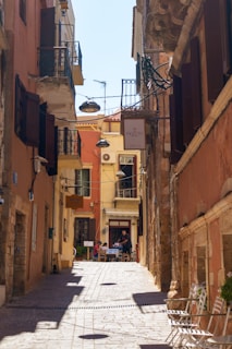 A narrow, charming alleyway in a Mediterranean town is lined with rustic buildings displaying warm earth tones. The cobblestone path leads to an outdoor cafe where people are sitting at tables enjoying a meal. Hanging lamps and signs add character, and balconies with wrought iron railings are seen on the upper floors.