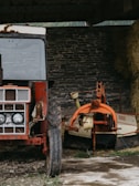 A rustic scene featuring an old, weathered red tractor with visible rust and scratches on its body. Beside the tractor is an orange and beige farm implement, possibly used for hay harvesting, with a large circular structure attached. The background consists of a rough, stone brick wall, and a stack of hay is partially visible to the right. The ground is partially covered with grass and dirt.