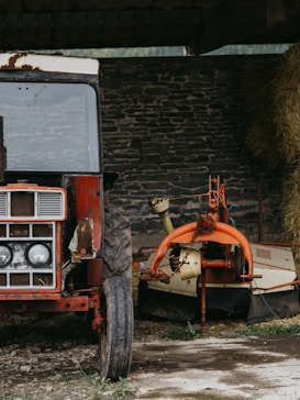 A rustic scene featuring an old, weathered red tractor with visible rust and scratches on its body. Beside the tractor is an orange and beige farm implement, possibly used for hay harvesting, with a large circular structure attached. The background consists of a rough, stone brick wall, and a stack of hay is partially visible to the right. The ground is partially covered with grass and dirt.