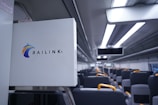 The interior of a modern train is depicted, featuring rows of seats with gray upholstery and yellow armrests. Above the seats are overhead compartments and sleek lighting fixtures. A sign with the Railink logo is visible in the foreground.