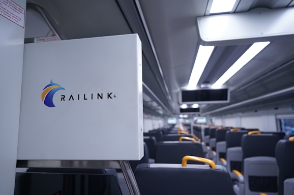The interior of a modern train is depicted, featuring rows of seats with gray upholstery and yellow armrests. Above the seats are overhead compartments and sleek lighting fixtures. A sign with the Railink logo is visible in the foreground.