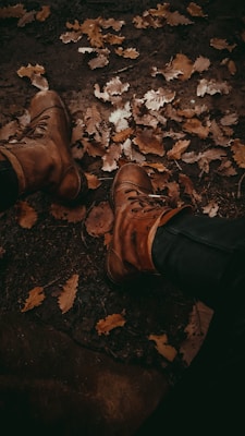 A pair of brown leather boots surrounded by fallen autumn leaves on a muddy ground. The leaves are in shades of brown, yellow, and orange, creating a rustic and natural setting.