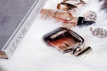 A collection of elegant items is placed on a soft white fur surface. There is a grey book titled 'Lean In' alongside two perfume bottles. A pair of intricate gold earrings and a bold gold cuff bracelet complement the luxurious display.