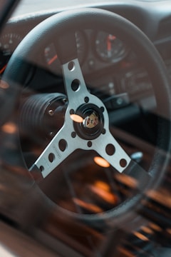 A close-up view of a car's steering wheel with a metallic finish, featuring a circular hub with multiple holes and a logo at the center. The dashboard and gauges are visible in the background through a slightly blurred windshield.