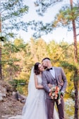A couple in formal wedding attire stands close together in a natural setting by a lake. The groom is wearing a light grey suit with a bow tie, while the bride is in a white lace gown with a veil. They are surrounded by tall trees with green foliage and the background shows sunlight filtering through the forest, creating a peaceful and romantic atmosphere. The groom holds a colorful bouquet featuring orange and white flowers.