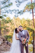A couple in formal wedding attire stands close together in a natural setting by a lake. The groom is wearing a light grey suit with a bow tie, while the bride is in a white lace gown with a veil. They are surrounded by tall trees with green foliage and the background shows sunlight filtering through the forest, creating a peaceful and romantic atmosphere. The groom holds a colorful bouquet featuring orange and white flowers.