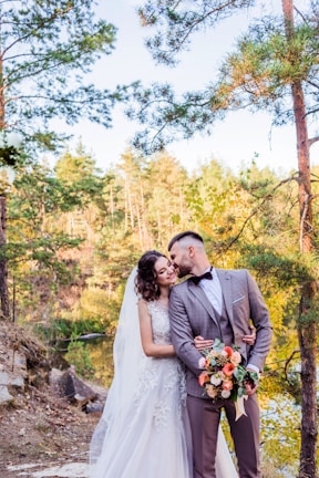 A couple in formal wedding attire stands close together in a natural setting by a lake. The groom is wearing a light grey suit with a bow tie, while the bride is in a white lace gown with a veil. They are surrounded by tall trees with green foliage and the background shows sunlight filtering through the forest, creating a peaceful and romantic atmosphere. The groom holds a colorful bouquet featuring orange and white flowers.