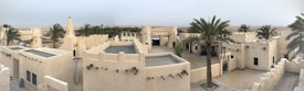 A panoramic view of a traditional desert village with beige, flat-roofed buildings featuring crenellated walls and small windows. Several tall palm trees are scattered throughout the area, adding greenery to the sandy environment. The horizon shows a vast desert landscape under a clear sky.