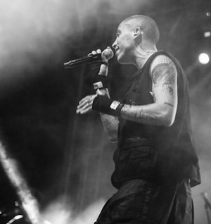A black-and-white image captures a person passionately performing on stage with a microphone in hand. The person has tattoos on their arm and is wearing gloves and a sleeveless top, as well as an earpiece. Smoke or mist swirls in the background, adding to the dynamic atmosphere.