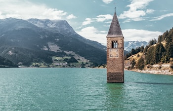 A lone bell tower emerges from the center of a serene, turquoise lake. Surrounding the lake, lush green hills and distant snow-capped mountains form a picturesque backdrop. The scene is tranquil and it evokes a sense of history and mystery.