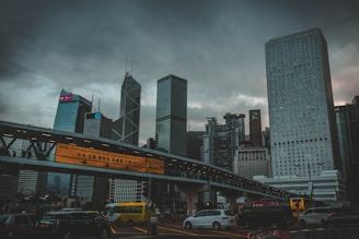 A cityscape featuring a skyline with modern skyscrapers under a cloudy sky. Notable architectural structures include buildings with unique patterns and designs. A pedestrian bridge with illuminated signage crosses above a busy urban road where various vehicles, including cars and buses, are traveling.