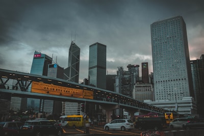 A cityscape featuring a skyline with modern skyscrapers under a cloudy sky. Notable architectural structures include buildings with unique patterns and designs. A pedestrian bridge with illuminated signage crosses above a busy urban road where various vehicles, including cars and buses, are traveling.