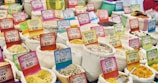 Numerous sacks filled with various types of grains, rice, and spices are displayed in a market setting. Each sack is accompanied by a colorful price tag indicating the product name and cost in euros. The arrangement appears vibrant and diverse, showing a rich assortment of culinary ingredients.