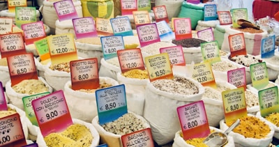 Numerous sacks filled with various types of grains, rice, and spices are displayed in a market setting. Each sack is accompanied by a colorful price tag indicating the product name and cost in euros. The arrangement appears vibrant and diverse, showing a rich assortment of culinary ingredients.