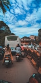 Motorcyclists navigate through a busy street alongside a stationary bus. The sky is partly cloudy with palm trees visible on the side. Several urban buildings are in the background, and there are barriers separating traffic lanes.