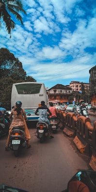 Motorcyclists navigate through a busy street alongside a stationary bus. The sky is partly cloudy with palm trees visible on the side. Several urban buildings are in the background, and there are barriers separating traffic lanes.