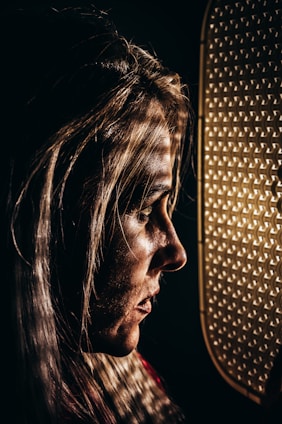 A profile view of a person with long, light-colored hair, interplaying with dramatic lighting casting shadows across the face. The person is looking towards a patterned surface illuminated with a warm glow.