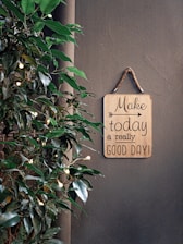 A motivational sign with the phrase 'Make today a really good day!' hangs on a rope against a plain wall. To the side, there is a plant with green leaves adorned with small glowing lights, adding a touch of warmth and life.