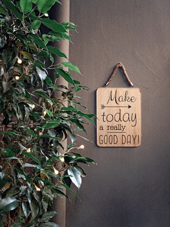 A motivational sign with the phrase 'Make today a really good day!' hangs on a rope against a plain wall. To the side, there is a plant with green leaves adorned with small glowing lights, adding a touch of warmth and life.